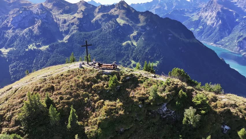Aerial view of a mountain summit featuring a large cross and people sitting on a wooden bench, surrounded by rugged alpine peaks and green slopes in Switzerland.