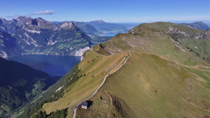 Aerial view of a winding path along a mountain ridge overlooking a deep blue lake surrounded by alpine peaks in Switzerland.