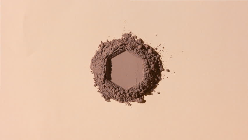 From-above view of a mountain of brown powder with a hexagonal engraving on a beige backdrop lit by orbiting light source projecting changing shadows