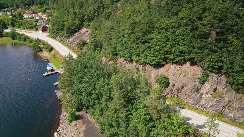 Aerial view of the scenic coastal road winding past rocky cliffs and dense forests near Sogne, featuring small boat docks along the water in Kristiansand, Agder, Norway.