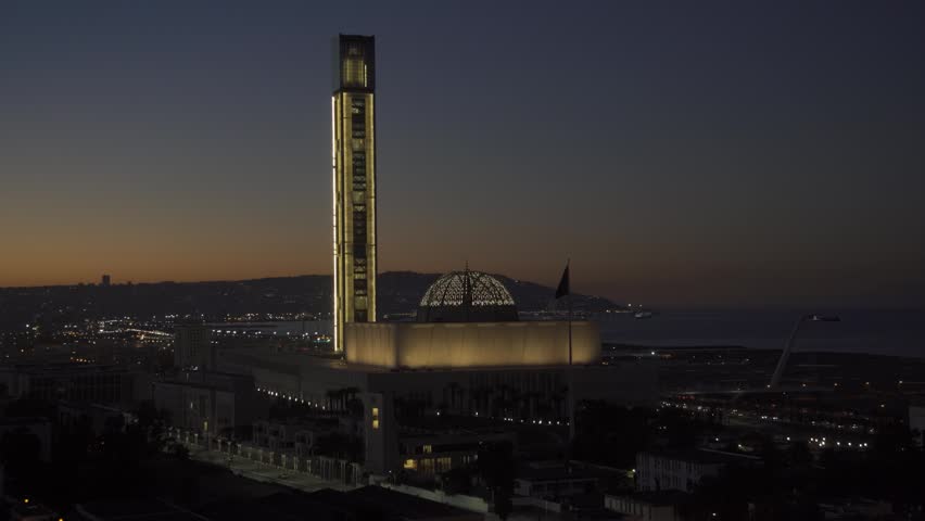 Aerial view of the illuminated Great Mosque of Algiers at night, featuring its tall minaret and dome against the coastal city lights in Algeria.