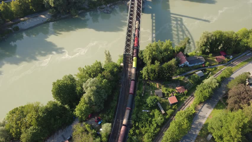 Aerial top down view of a freight train crossing the railway bridge over the Inn river toward Passau, Lower Bavaria, Germany, filmed on 17 July 2026. The drone looks straight down as the train moves across the bridge, with the shadow of the steel truss construction cast on the water. Footage of rail freight transport and logistics.
