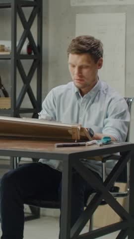 Focused young architect working at drafting table in modern studio interior, sketching detailed building plans and technical drawings with professional precision