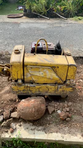 A yellow vibratory plate compactor machine parked on the dirt ground next to an asphalt road under construction.