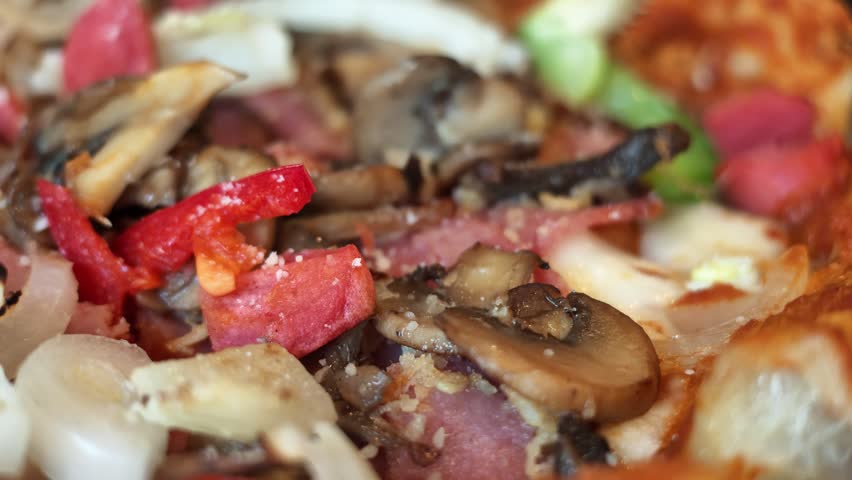 Macro slow motion view panning across rich toppings of sliced mushrooms, red bell peppers, onions, and melted cheese on hot pizza.
