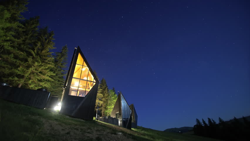 Modern angular cabins stand beneath a clear star filled night sky, Exterior lights illuminate dark facades and pine forest on a grassy slope