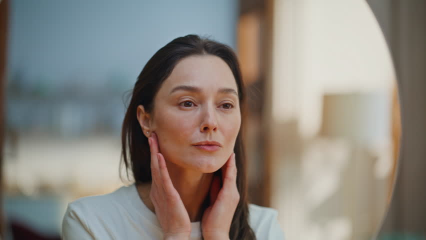 Woman examining face in mirror searching wrinkles aging signs closeup. Serene brunette carefully studying skin texture at daily skincare ritual. Smiling lady enjoy reflection satisfied natural beauty.