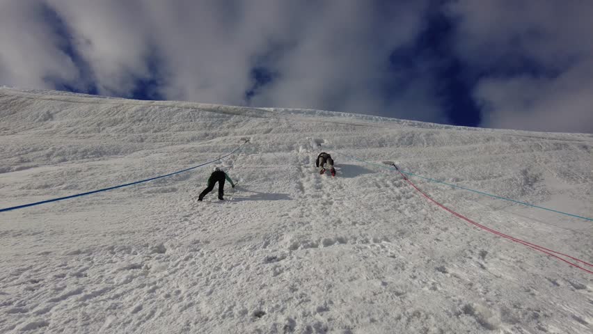 Mountaineers Climbing Steep Snow Slope in Antarctica - Two climbers scale a textured ice wall with safety ropes under a cloudy sky.