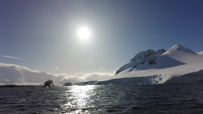 Antarctic Expedition Ship Near Snow-Covered Mountains - Vessel navigates icy waters beneath bright sun and massive polar peaks. High-contrast landscape featuring glaciers and choppy seas.