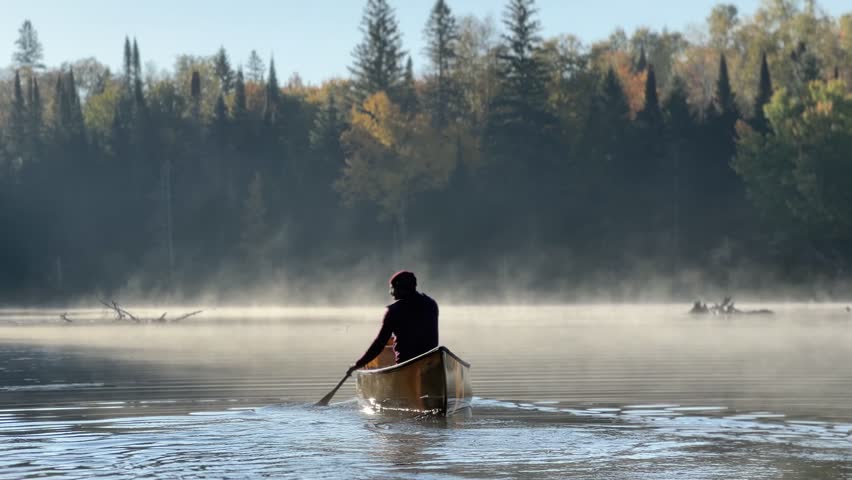 A Hispanic man paddling solo across a misty lake in Algonquin Park, Ontario. Morning mist and calm water reflect the peaceful wilderness scene