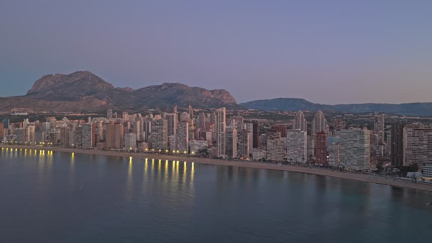 Aerial footage capturing the stunning benidorm cityscape and its iconic beach at dusk. The vibrant city lights reflect on the calm sea