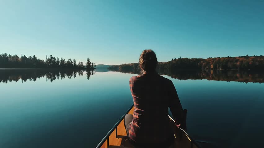 A Caucasian woman at the bow paddling across a crystal-clear, still lake in the backcountry, with forest in the background in Algonquin Park, Ontario