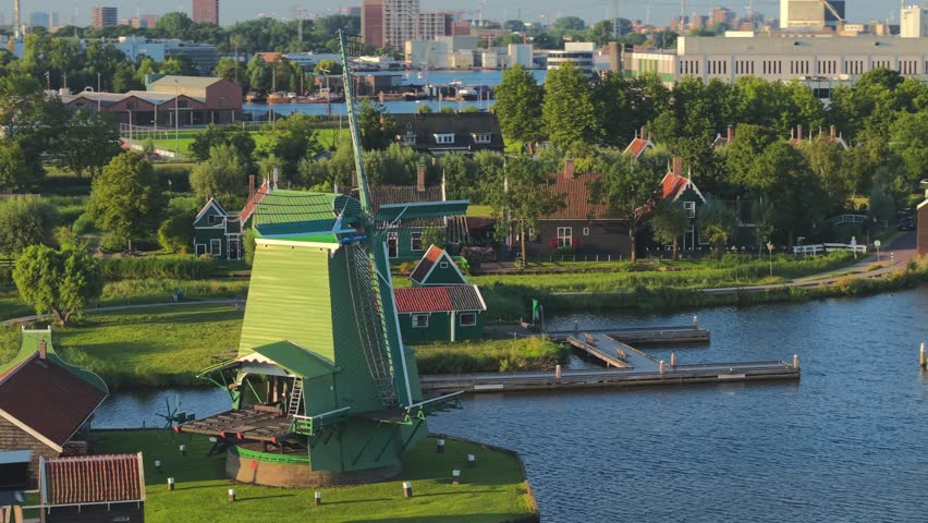 A Drone circling a green windmill by the canal with houses, farms and open meadows nearby in Netherlands
