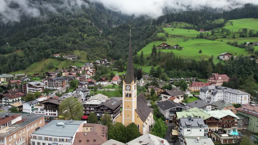 Aerial view of the historic church in Bad Gastein surrounded by traditional alpine architecture, lush green hills, and misty mountains in Bad Gastein, Salzburg, Austria.