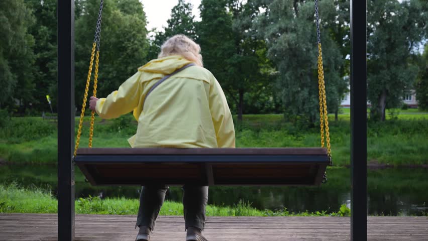 Woman sits alone on park swing. Swings gently above grassy pond. Trees frame the peaceful scene. Soft light falls on his yellow coat. No animals or plants visible. Quiet moment in public park