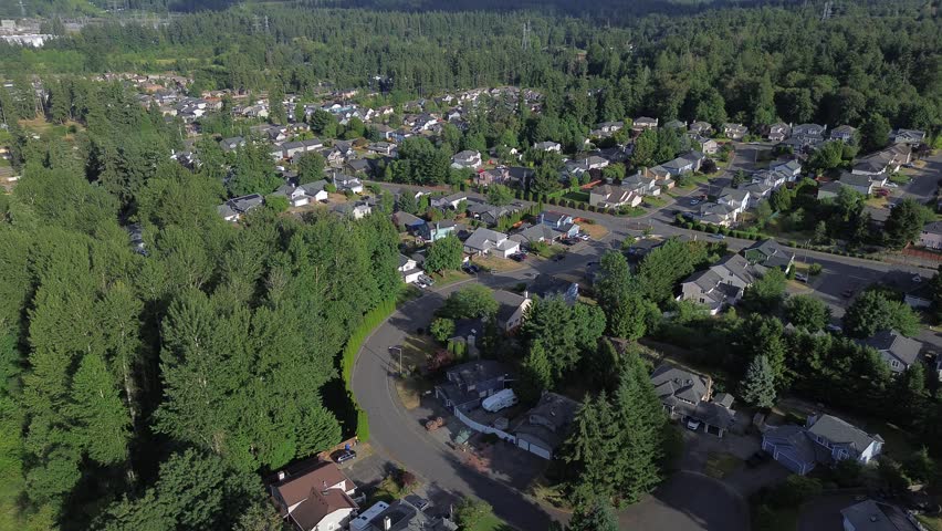 Meandering pavement tracks through residential belts bordered by tall, evergreens throughout Kent, WA. Aerial perspective reveals nature of neighborhood pathways, where daily life remains. USA