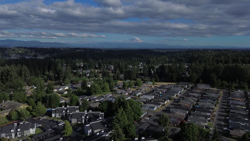 Mount Rainier peaks above forests, framing residential zones where streets sprawl across Kent, WA. Low hanging clouds drift over evergreen canopy, uniting neighborhoods with glacial topography. USA