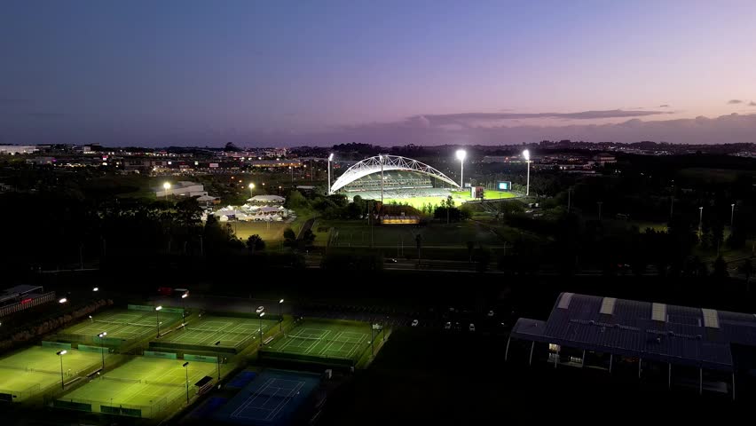 An aerial shot of a lighted baseball stadium and tennis courts surrounded by Albany suburban area under a purple and orange sunset sky in New Zealand