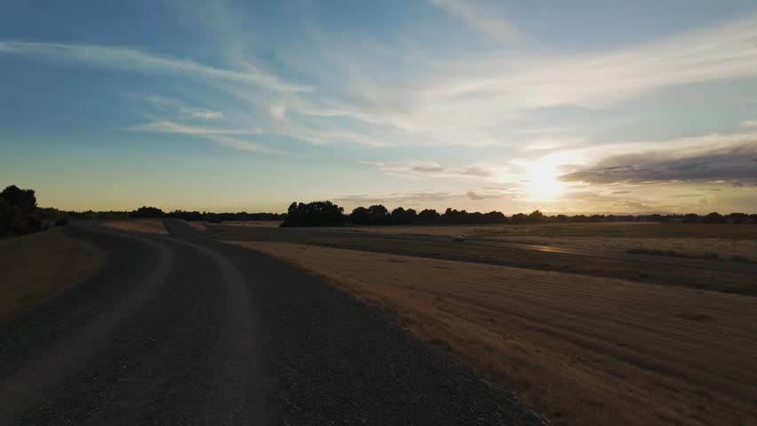 A winding gravel road curves through golden fields under a glowing sunset sky with streaked clouds