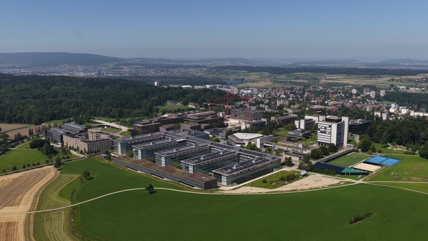 An aerial view of HCP ETH Honggerberg campus surrounded by green fields and a forest, with a town and hills in the background in Zurich, Switzerland