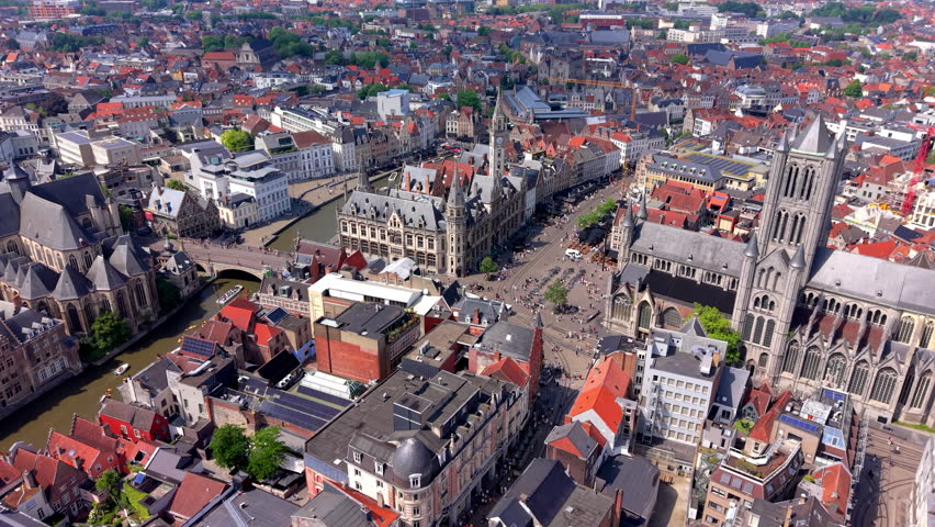 A magnificent wide-angle aerial photograph capturing a high-altitude drone overview of the stunning architectural heritage in the historic center of Ghent, Belgium. The perspective highlights the massive gothic stone architecture of Saint Nicholas Church (Sint-Niklaaskerk) rising prominently on the right. In the center, the ornate towers and clock spire of the historic Old Post Office (Voormalig Postkantoor) building overlook the vibrant Korenmarkt square, which is filled with crowds of walking pedestrians and tram tracks. To the left, a scenic river canal flows past traditional brick guildhouses, crossed by the historic Saint Michael