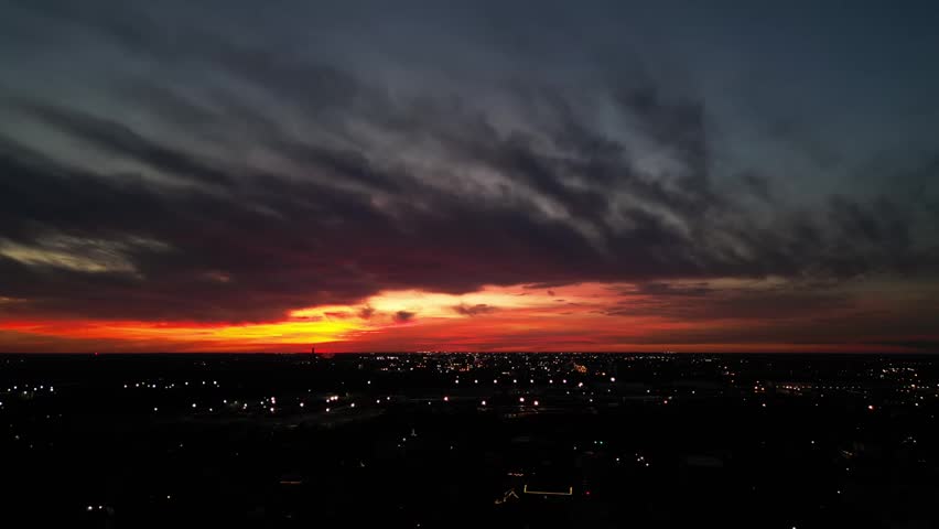 An aerial view of a dramatic sunset over the Indianapolis skyline, with streetlights beginning to glow in the darkness below a vibrant, colorful sky