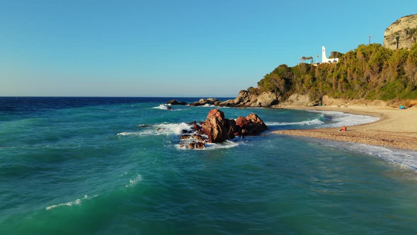 An aerial view of a white chapel on Karlovasi with an exotic rocky beach and wooded cliff under blue sky on the island of Samos, North Aegean, Greece