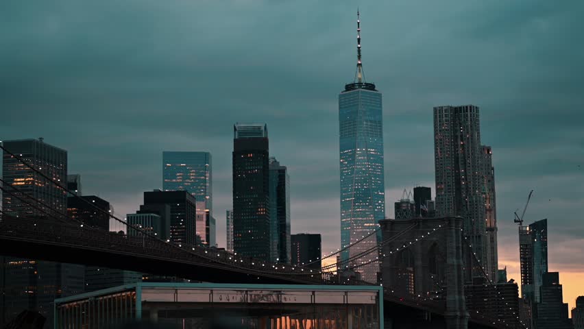 A sunset scene of the New York City skyline, featuring the Freedom Tower and the Brooklyn Bridge in the foreground, all under a dramatic cloudy sky.