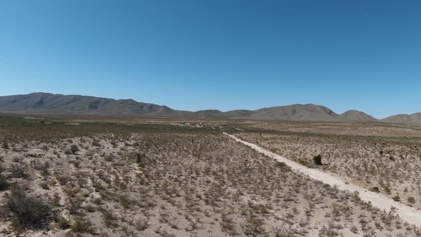 A low drone view of a dirt road cuts through a dry desert landscape with low-lying brush and small plants, and hills in distance on a sunny clear day