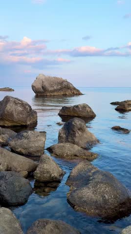 Vertical 9:16 locked‑off shot. Rocky shoreline with large boulders in clear shallow water