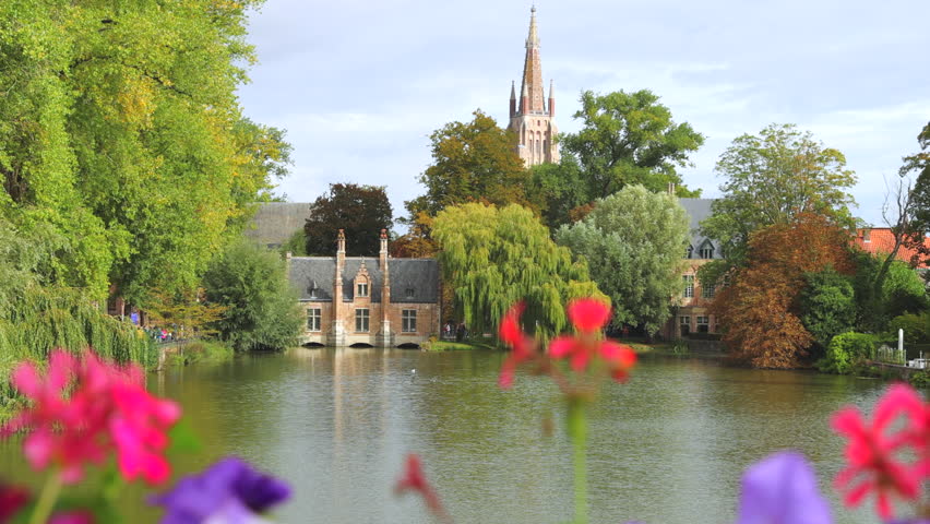 Bruges town in Belgium, Europe. Minnewater Lake Serenity: Beguinage and Church Spire Framed by Lush Greenery and Vibrant Flowers on a Calm Autumn Day