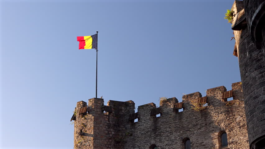 Waving Belgium Flag Over Old Stone Castle Wall with Battlements and Turrets Under Clear Blue Sky on Sunny Day. Gravensteen, a medieval castle in the city of Ghent in Belgium, Europe.