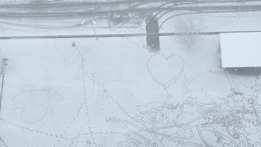 Aerial top-down view of a little girl in a red jacket walking and playing among heart-shaped footprints in deep snow during a blizzard.