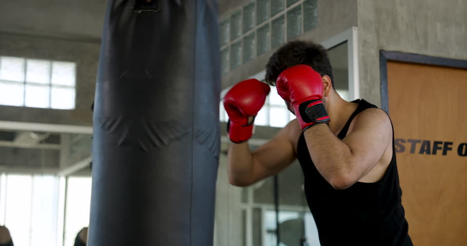 Adult Caucasian man strikes heavy bag with red boxing gloves in modern gym, alternating guard and punches. Male athlete builds power, endurance, coordination, confidence, active workout routine.