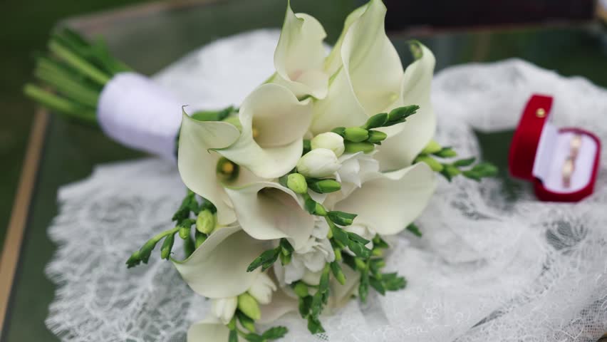 close-up of the wedding rings of the bride and groom and bouquet of pristine white calla lilies before the outdoor ceremony lie on the table.