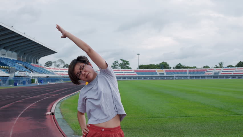 Authentic shot of an active Asian woman in sportswear stretching her arms on a running track. An empty stadium, green soccer field, and waving yellow flag provide excellent copy space.