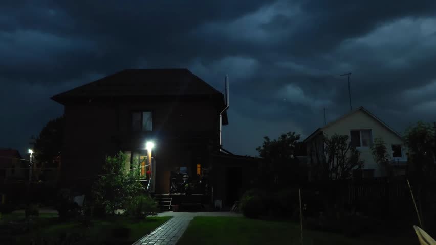 Exterior of residential house at night with bright illumination. Dusk view of cottage with glowing porch light under cloudy sky. Garden path in dark evening.