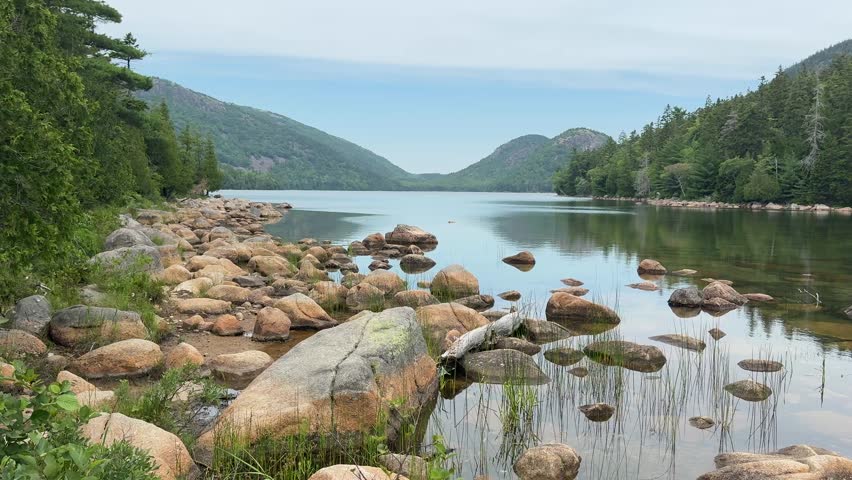 Wide scenic view of Jordan Pond in Acadia National Park, Maine, with calm reflective water, granite boulders, forested shoreline, and the Bubbles mountains in the distance.