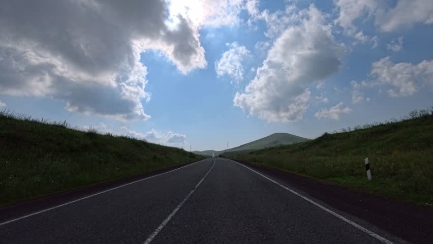 Long straight road stretches under bright blue sky with fluffy white clouds. Sun shines brightly above horizon. Green hills roll gently on both sides