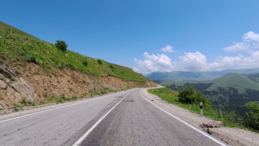 Mountain road curves through green hills under bright blue sky. Clouds float above distant peaks. Road edges marked with white lines