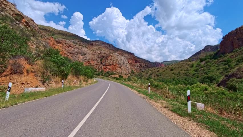 Road winds through red rock canyon under blue sky. Green trees line both sides of asphalt path. Sunlight casts shadows on road surface