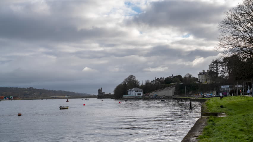 Two people row a boat on a river with a castle in the background on a cloudy day.