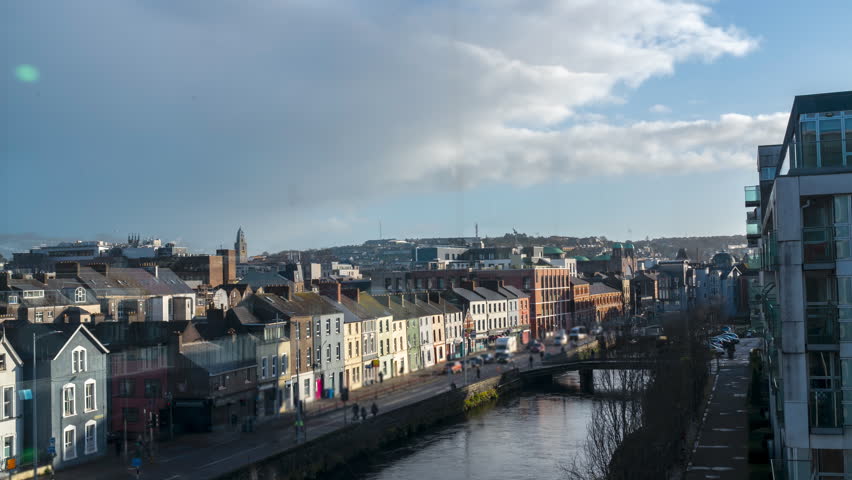 A view of the city of Cork, Ireland, with the River Lee flowing through it. The city is bustling with traffic and pedestrians, and the buildings are a mix of old and new architecture.