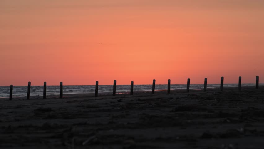 Louisiana, USA – sunset over the Gulf of Mexico coast. A tranquil coastal scene captured under a clear sky during the calm period often referred to as the "calm after sunset" or the "last light of day." A line of wooden pilings extends across the water on the horizon, a characteristic feature of many Gulf Coast beaches.