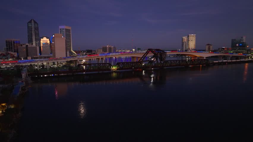 Cinematic drone footage of the Acosta Bridge crossing the St. Johns River in downtown Jacksonville, Florida. The video features the illuminated skyline, historic railroad bridge, and reflections on calm water during blue hour.