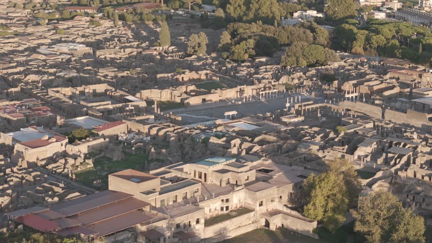 An aerial view captures the sunny vast, preserved ruins of the ancient city of Pompeii with stone walls and an uncovered forum with trees in Italy