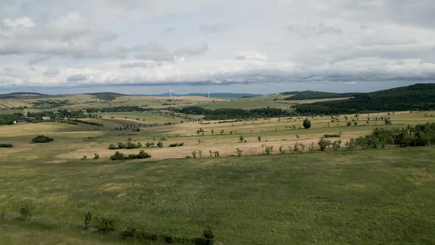Aerial shot of mountain plateau of the Ore Mountains (Krušné hory) under a dramatic, cloudy sky. Vast green and yellow agricultural fields spread across the landscape, with modern wind turbines