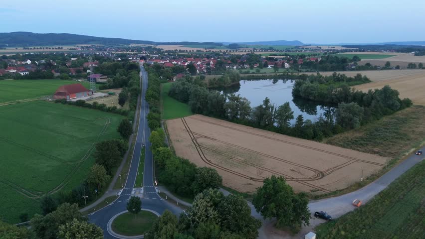 Aerial view of a small lake and a rural village beside a road with farmland and distant hills