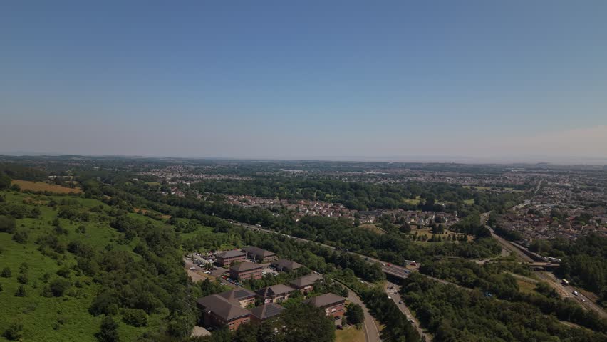 High-altitude aerial drone view of the M4 motorway near Cardiff, South Wales, United Kingdom. The footage captures moving traffic, surrounding residential neighbourhoods, green woodland and suburban development, highlighting the transport infrastructure connecting Cardiff with the rest of Wales. Ideal for transportation, commuting, infrastructure and urban planning concepts.