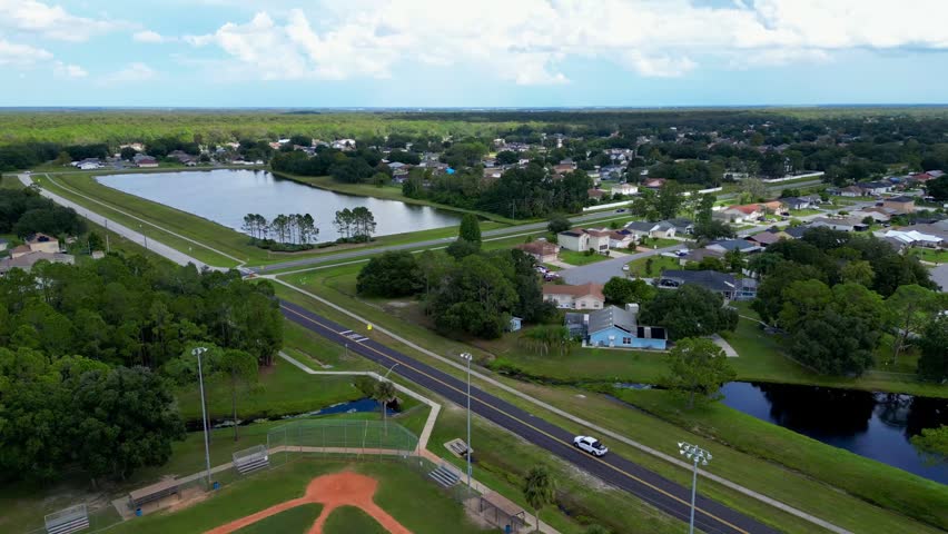 An aerial view of suburban area featuring a baseball diamond, road, and a large retention pond surrounded by houses and greenery in Poinciana, Florida